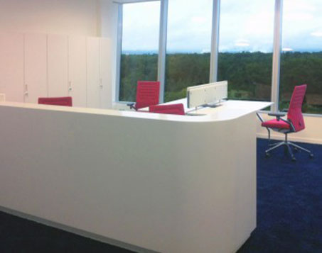 Bild des Empfangsbereichs des Bürogebäudes Squaire Frankfurt. Ein weißer Counter und rote Stühle, sind zu sehen.
